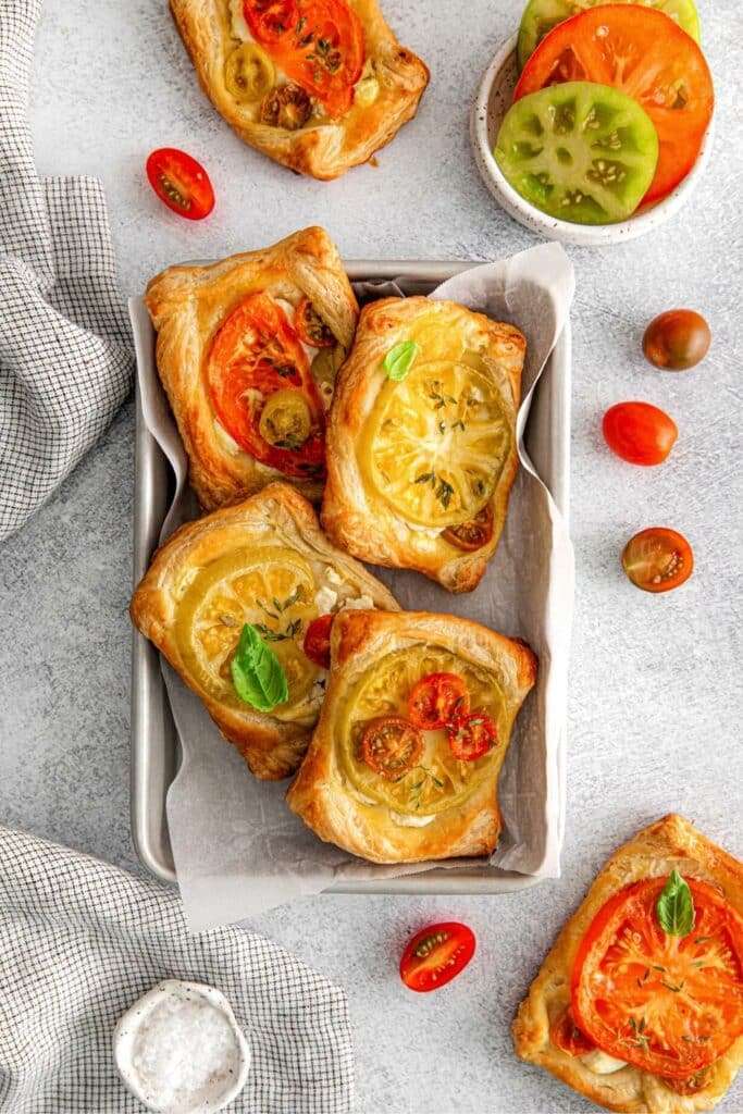 Four puff pastry tomato tarts on a metal serving tray next to sliced heirloom tomatoes.