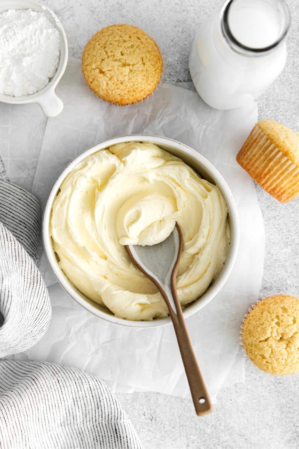 Best vanilla buttercream frosting in a bowl with spoon next to 3 unfrosted vanilla cupcakes.