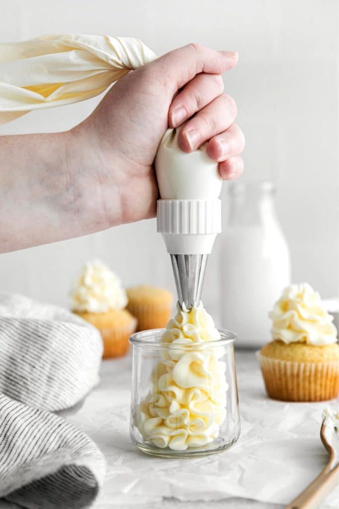 Hand squeezing vanilla frosting from a piping bag with metal tip into a small glass jar.