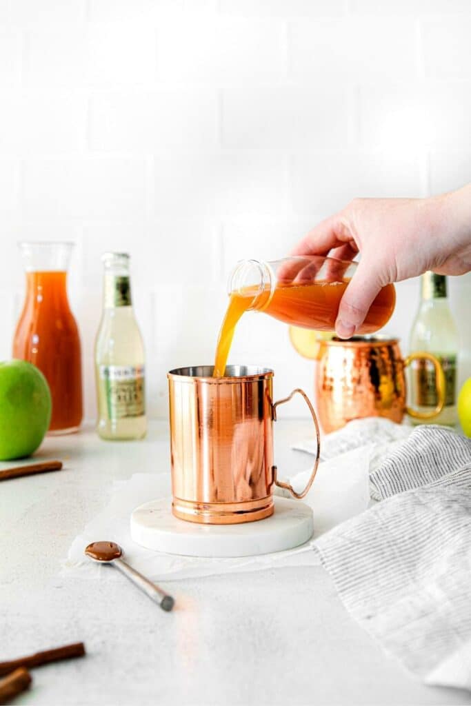 Fall Moscow mule being assembled on a white countertop with freshly-pressed apple cider.