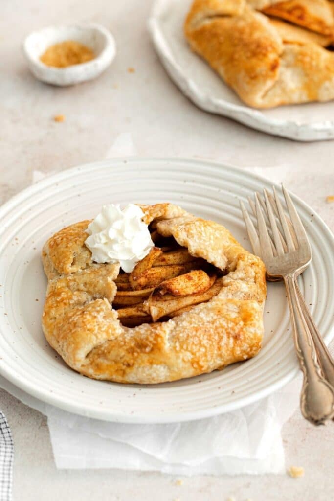 Individual-size apple galette with dollop of whipped cream on a white dessert plate with 2 sterling silver forks.