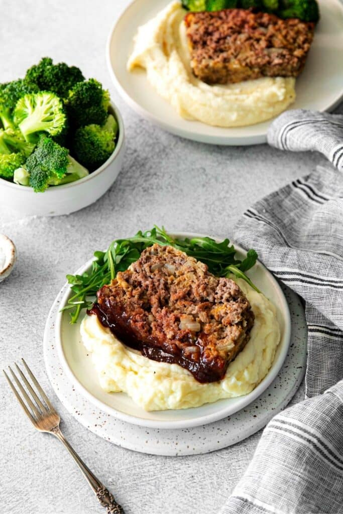 Dinner plate with BBQ meatloaf, mashed potatoes and salad next to a bowl of broccoli.