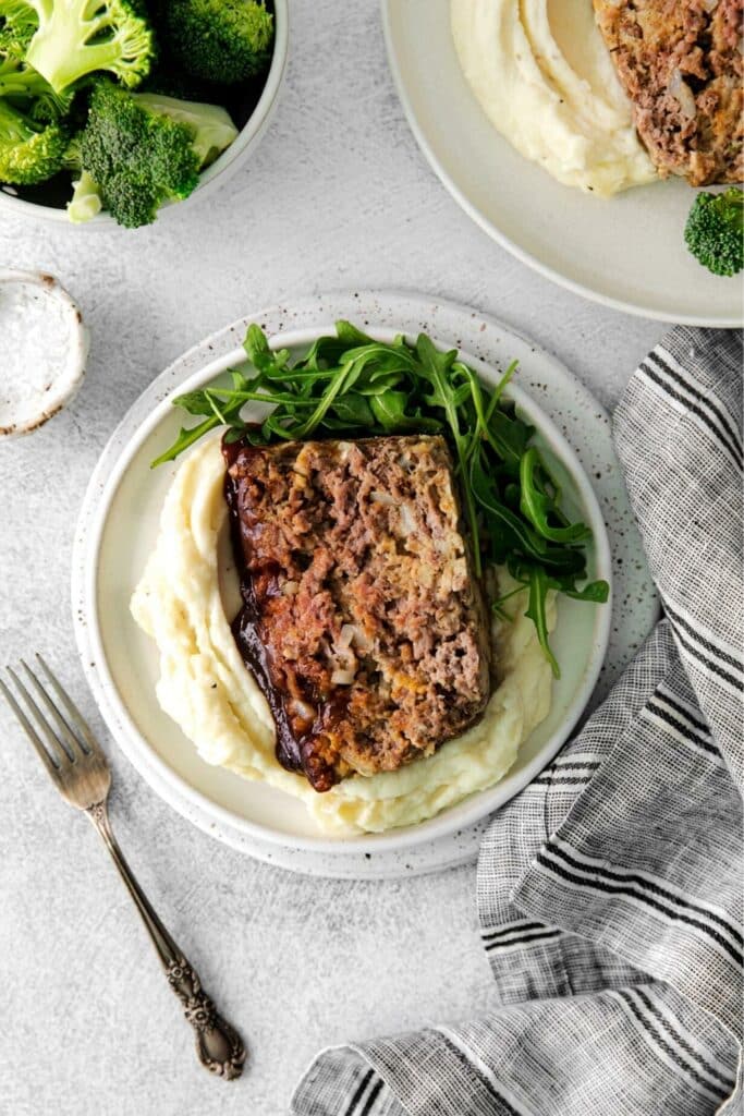 Slice of BBQ meatloaf topped with a savory meatloaf glaze plated with mashed potatoes and arugula.
