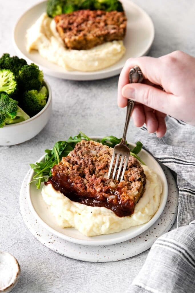 With fork in hand, digging into a plate of Southern BBQ Meatloaf on a dinner plate.