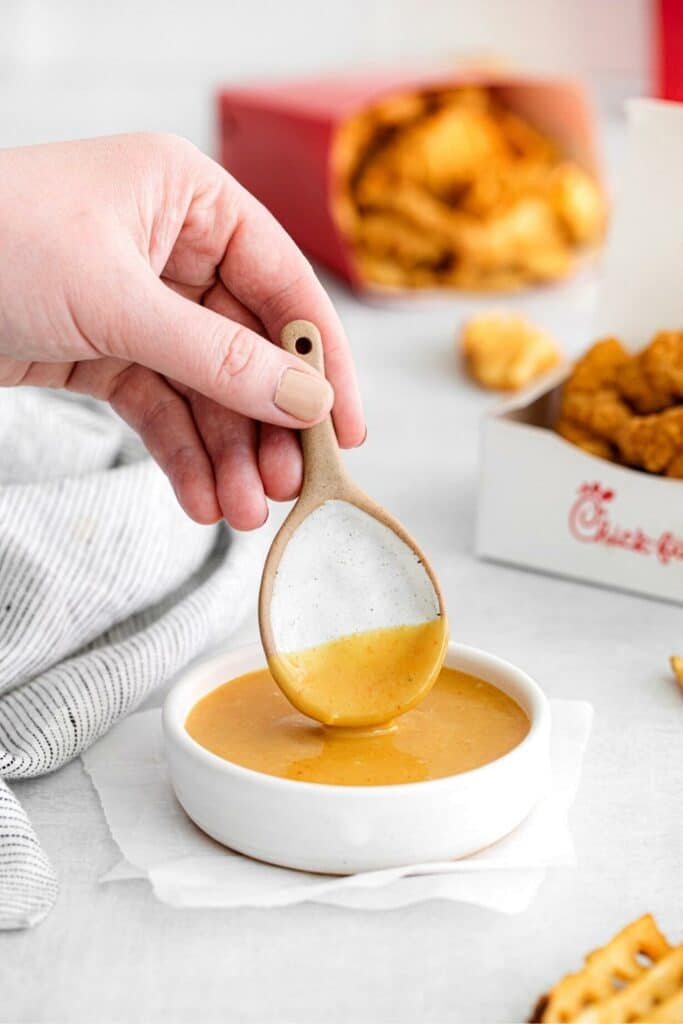 Hand with spoon lifting CFA dipping sauce up from a small white bowl. There's CFA food in the background.