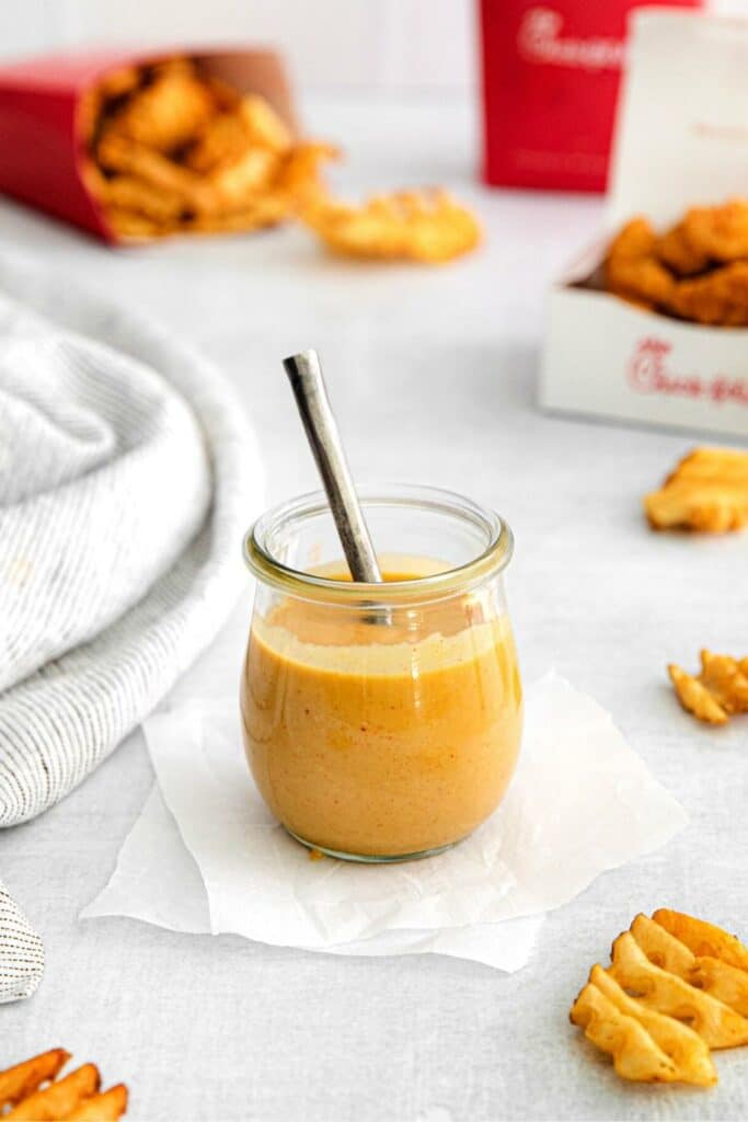 Homemade Chick-Fil-A sauce in a small glass jar with metal spoon next to CFA waffle fries and chicken nuggets.