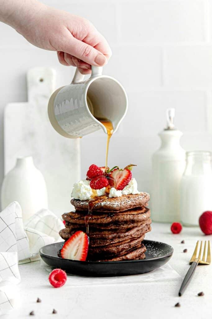 Hand holding a small ceramic pitcher and pouring maple syrup over a stack of chocolate pancakes.