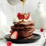 Maple syrup being poured over a stack of 5 chocolate pancakes garnished with mini chocolate chips, fresh strawberries, whipped cream.