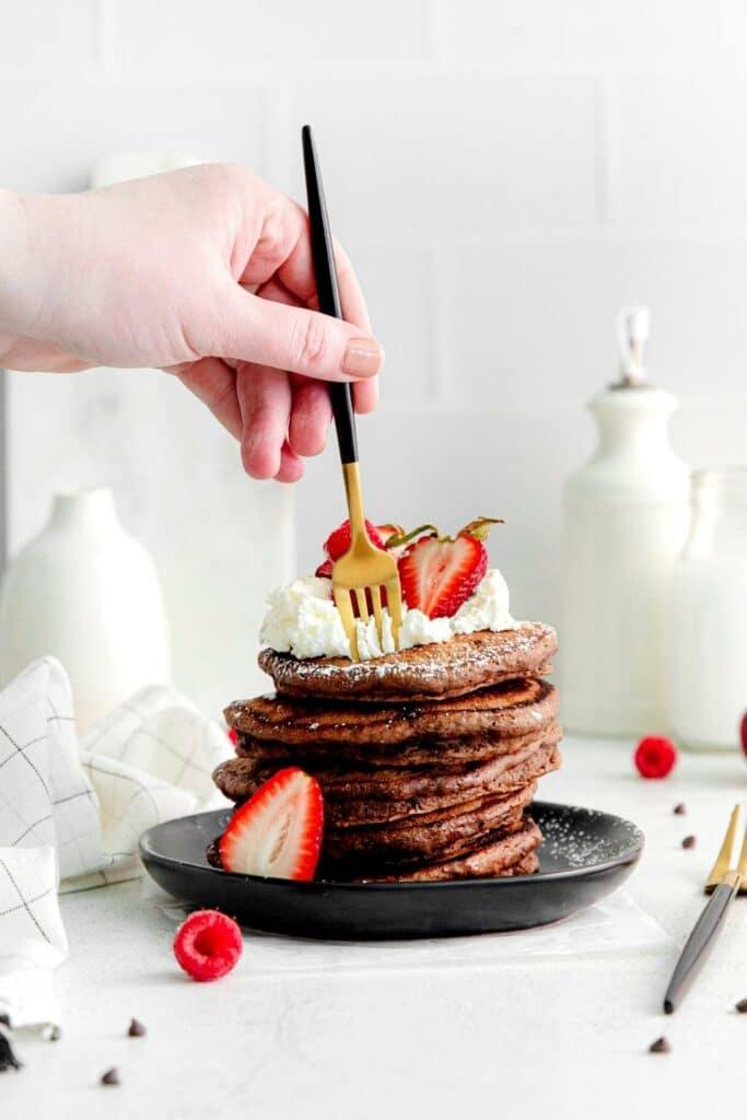 Hand with fork digging into a stack of chocolate pancakes garnished with whipped cream and strawberries.