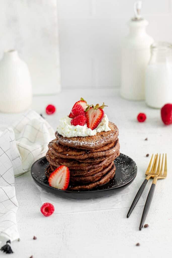 A tall stack of chocolate pancakes on a serving table with 2 black/gold forks.