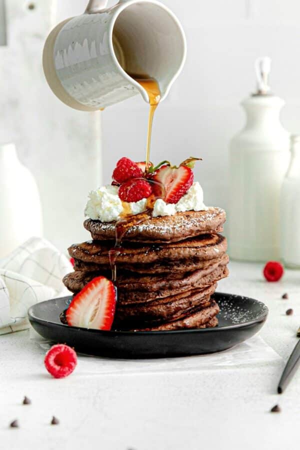 Maple syrup being poured over a stack of 5 chocolate pancakes garnished with mini chocolate chips, fresh strawberries, whipped cream.