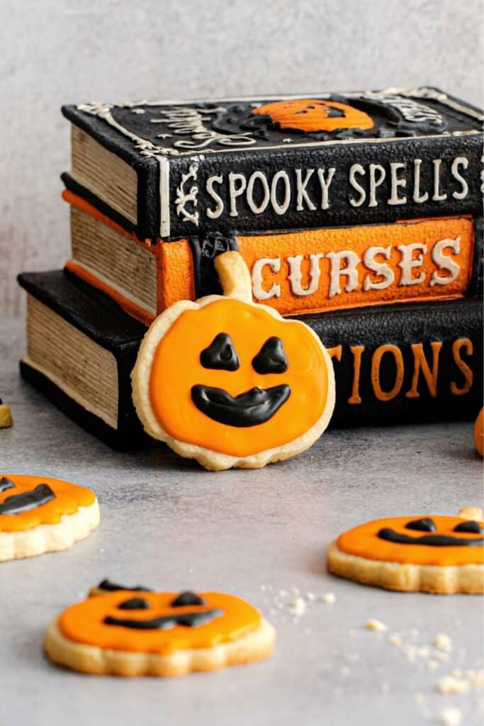 Halloween Jack-o'-lantern cookie leaning against a stack of books (spooky spells, curses, concoctions).