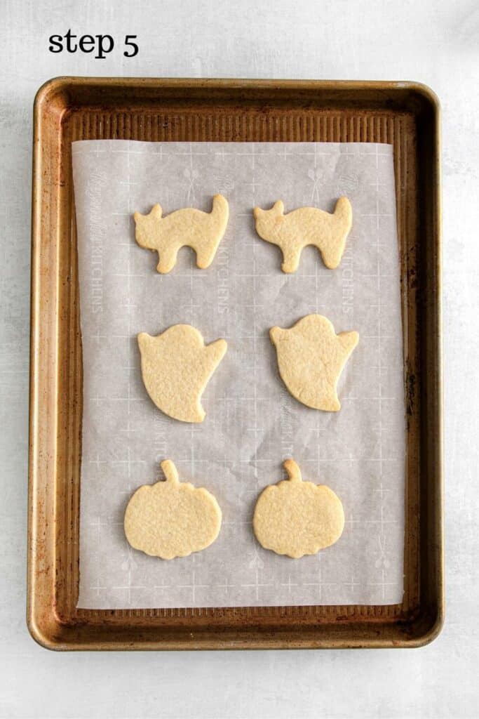 Six Halloween sugar cookies on a lined baking tray: 2 cats, 2 ghosts, 2 pumpkins.