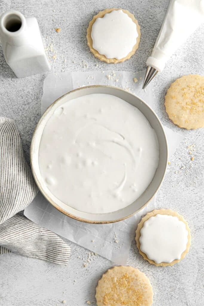 Bowl of royal icing next to frosted sugar cookies and a piping bag with metal tip.