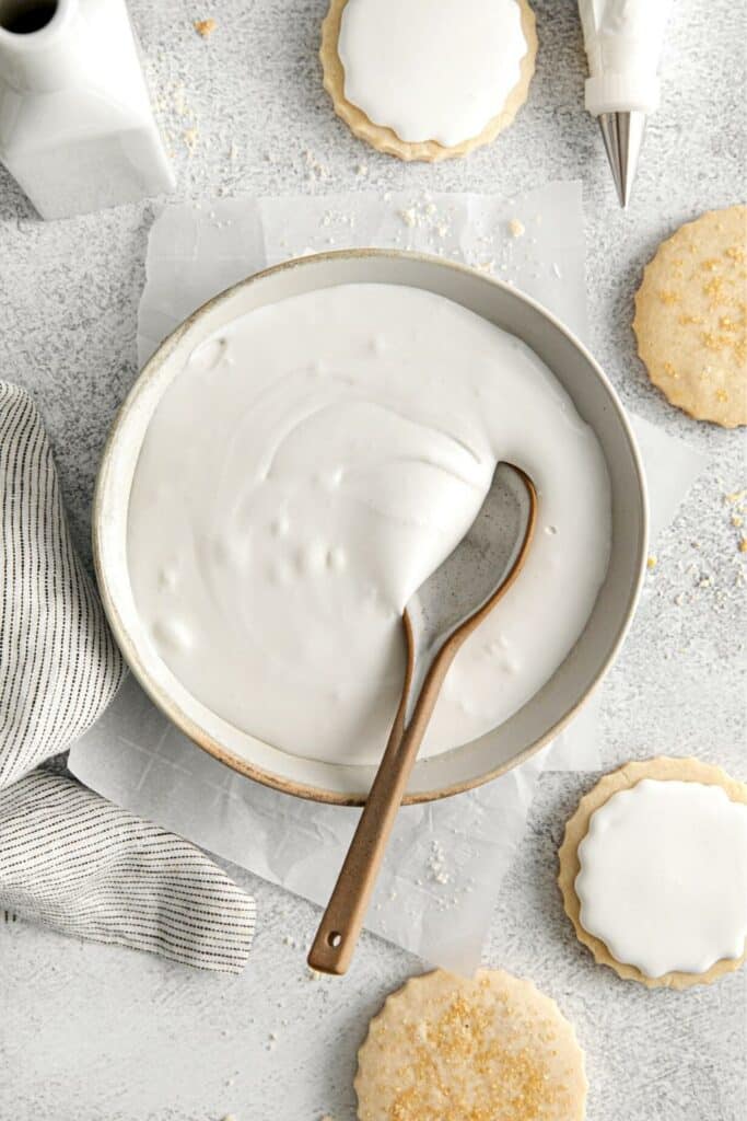 Royal icing in a bowl with spoon next to a piping bag with metal tip and frosted sugar cookies.