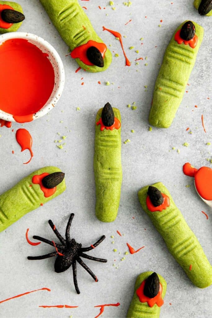 Green witch fingers cookies next to a black spider and a bowl of blood-red icing.