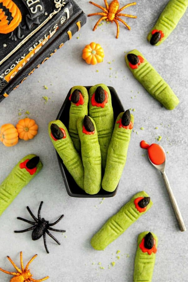 Halloween witch finger cookies served in a black coffin serving tray next to spooky spiders, pumpkins and spell books.