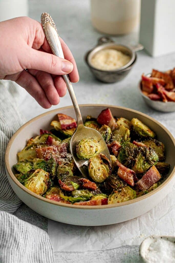 Hand with serving spoon scooping out Brussels sprouts with bacon from a serving bowl.