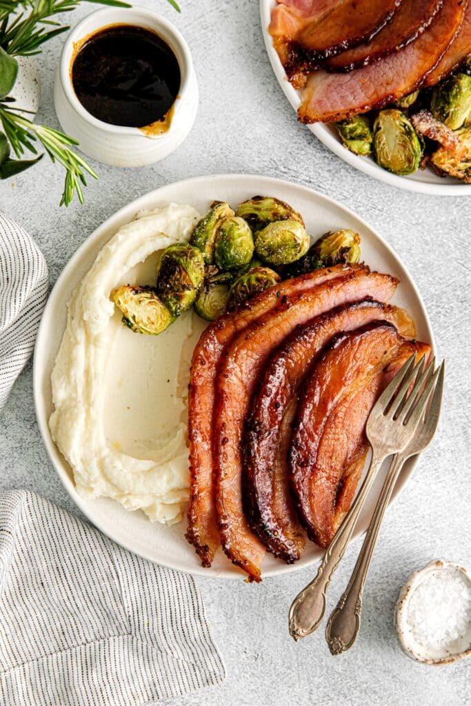 Slices of brown sugar glazed ham on a dinner plate with mashed potatoes and Brussels sprouts.