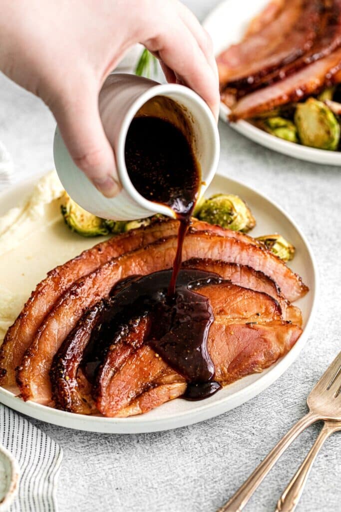Thick brown sugar glaze being poured over ham slices on a dinner plate.