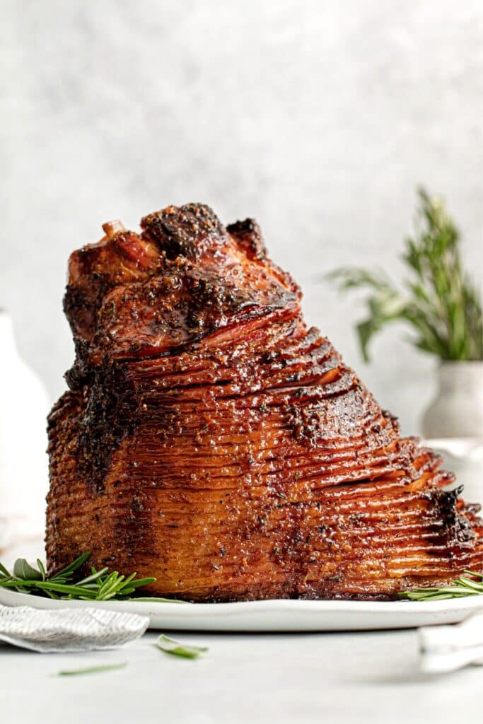 Spiral ham with brown sugar glaze on a white platter with fresh rosemary and sage.