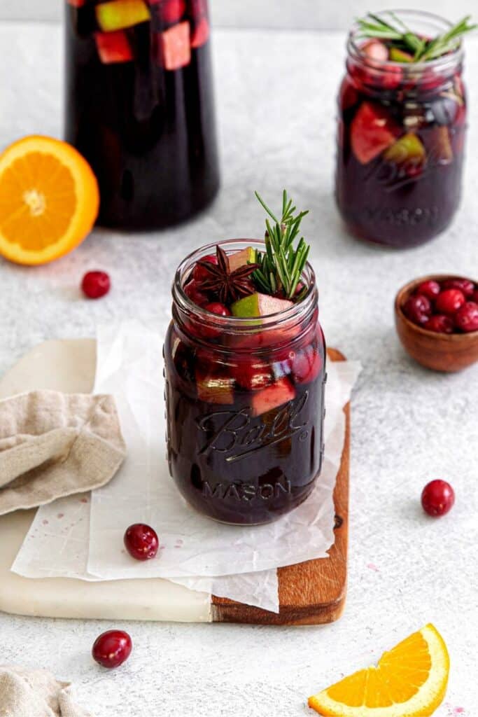 Holiday sangria in a tall glass pitcher with 2 mason jars of the drink garnished with rosemary.