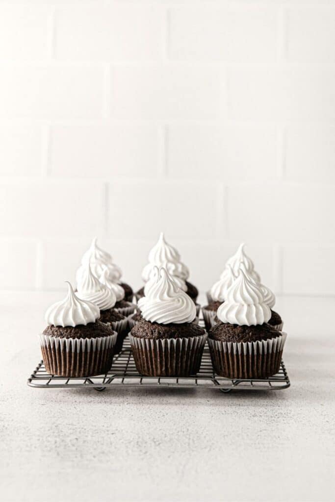 Swirls of marshmallow frosting on 12 chocolate cupcakes sitting on a wire rack.