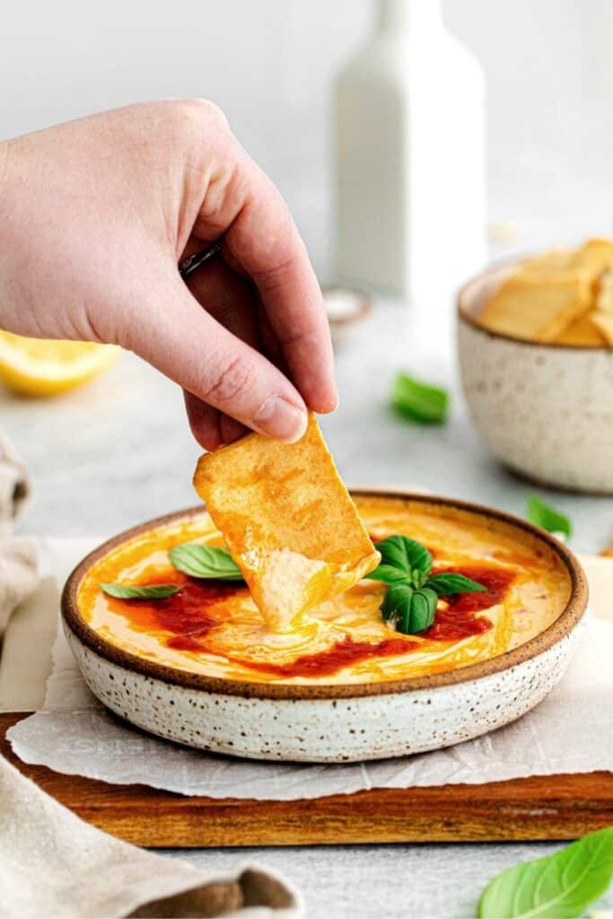 A hand dipping a pita chip into a cheesy spicy dip.