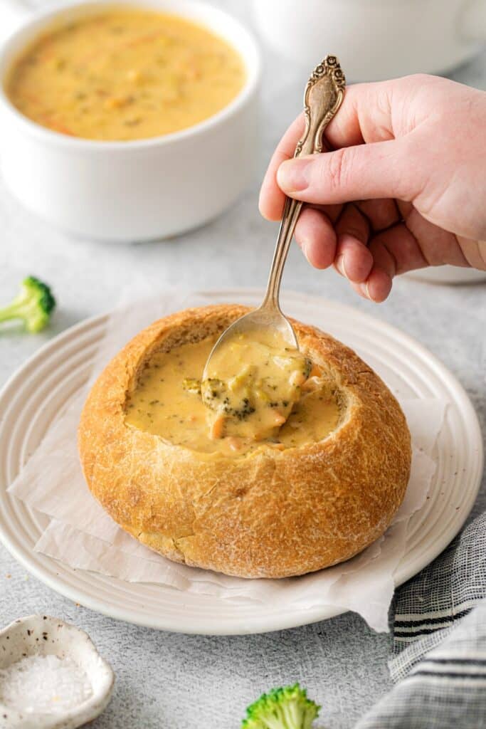 Hand lifting a spoonful of broccoli cheddar soup from a bread bowl.