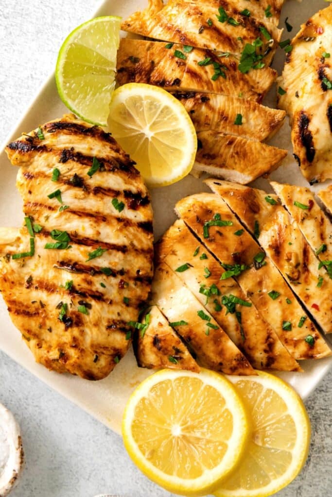 Sliced grilled chicken breasts on a platter with some ingredients from the Mexican citrus chicken marinade.
