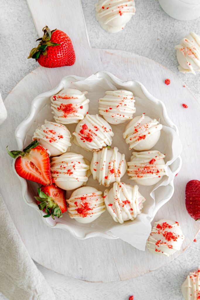 Ten strawberry truffles (cake balls) in a small white fluted cake dish along with fresh strawberries.