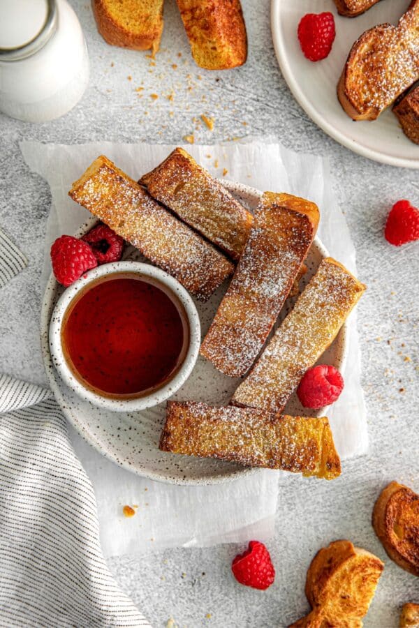 Air fryer French toast sticks plated and served with a ramekin of maple syrup and raspberries.