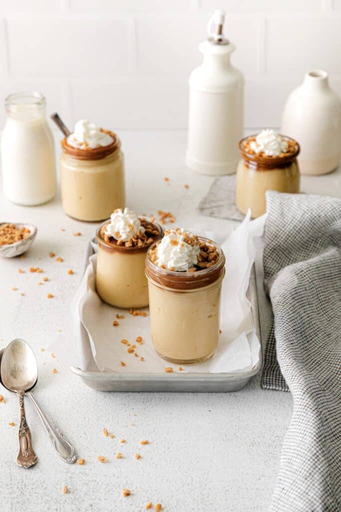 Four servings of butterscotch pudding garnished with whipped cream and nuts in small glass jars.