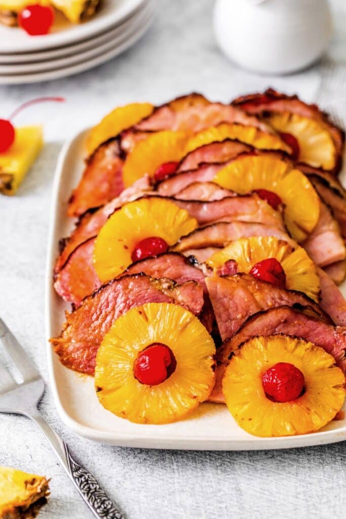 Thin slices of brown sugar pineapple ham on a serving tray with pineapple slices and maraschino cherries.