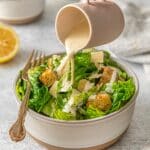 Creamy, healthy Caesar dressing being poured over Caesar salad - romaine lettuce, croutons, and shaved parmesan cheese.