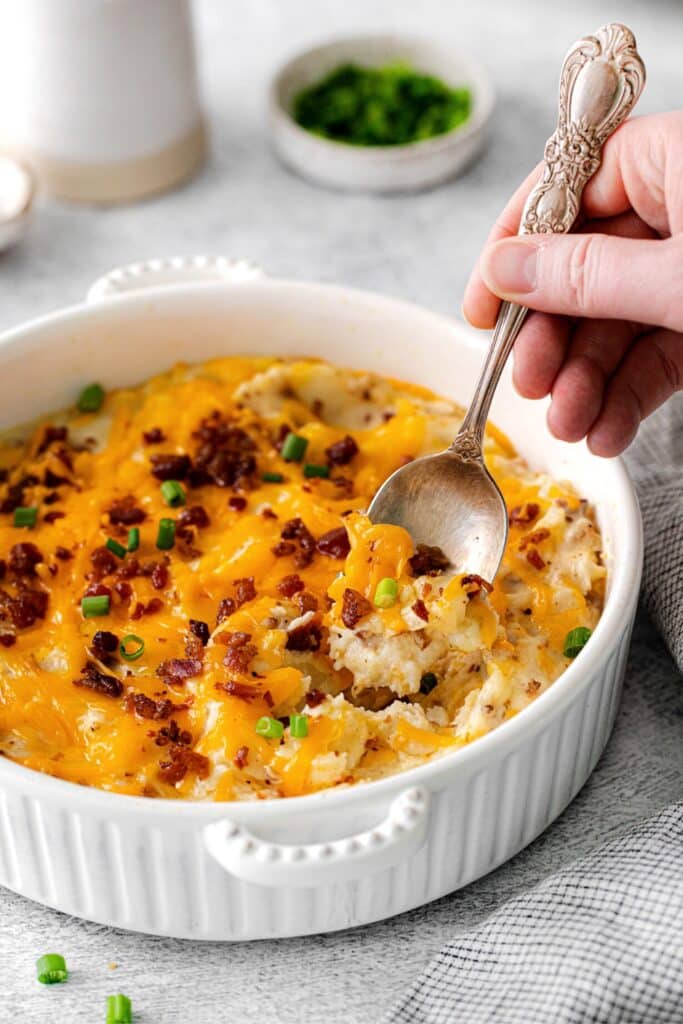 A serving spoon digging into a dish of loaded baked potato casserole topped with melted cheese.