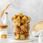Mini sourdough croutons in a glass jar next to a loaf of sourdough bread.