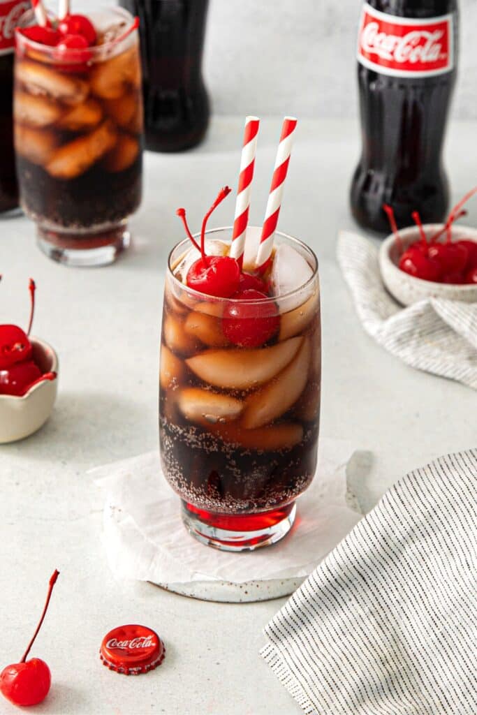 A Coca-Cola mocktail garnished with maraschino cherries and striped paper straws.