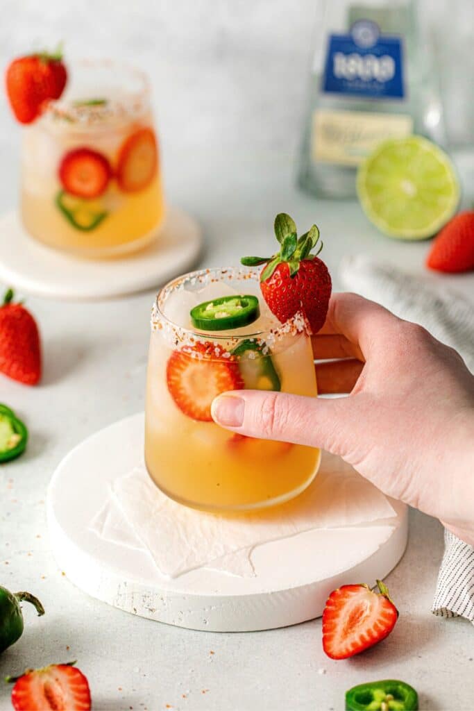 A hand lifting a cocktail glass of spicy strawberry jalapeño Margarita.