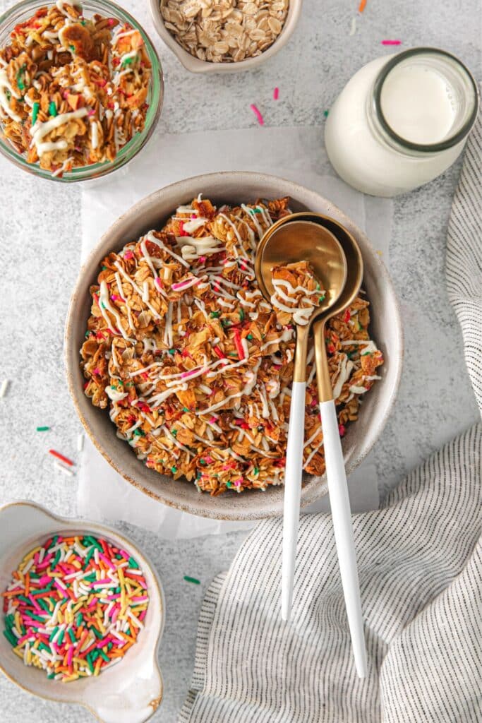 Birthday cake granola in a bowl with 2 spoons next to funfetti sprinkles and a glass bottle of milk.