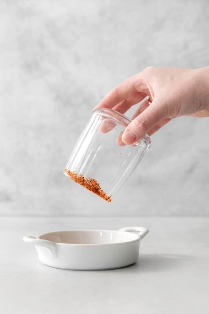 Dipping the rim of a glass into a small dish that has Tajin seasoning.