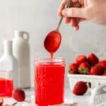 Homemade strawberry glaze dripping off a spoon into a mason jar next to fresh strawberries.