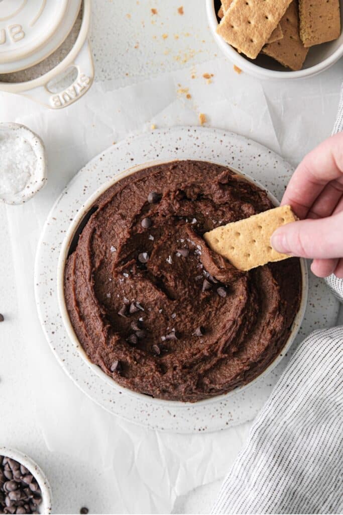 A hand holding a graham cracker, dipping it into a bowl of dark chocolate hummus.