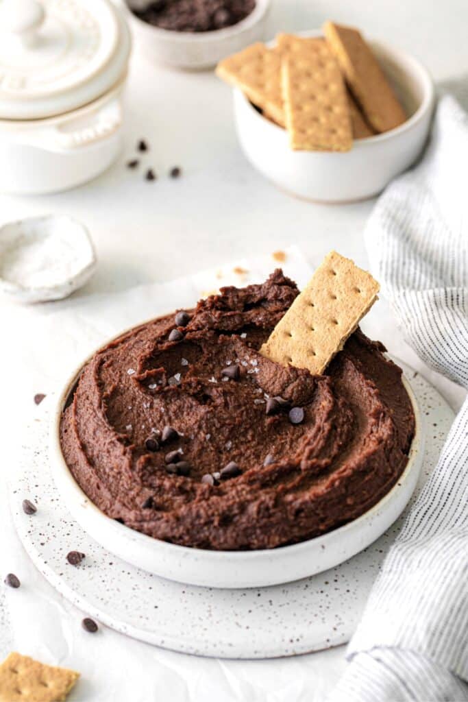 Bowl of dark chocolate hummus on a serving table with graham crackers and a pinch bowl of flaky salt.