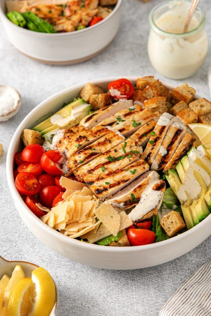 A freshly-assembled grilled chicken Caesar salad in a white serving bowl with Caesar salad dressing.