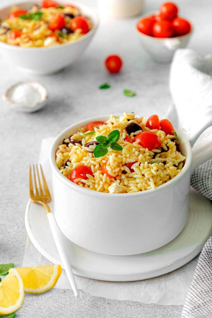 A single serving of summer orzo salad in a white cup with a gold-and-white fork.