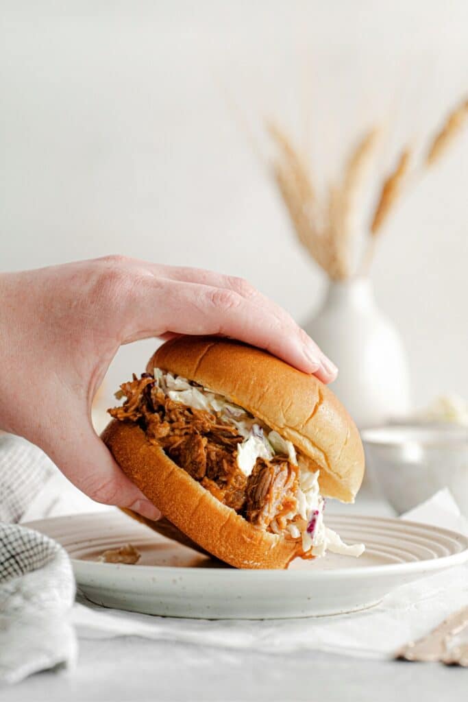 A hand reaching and lifting a pulled pork sandwich off a plate.