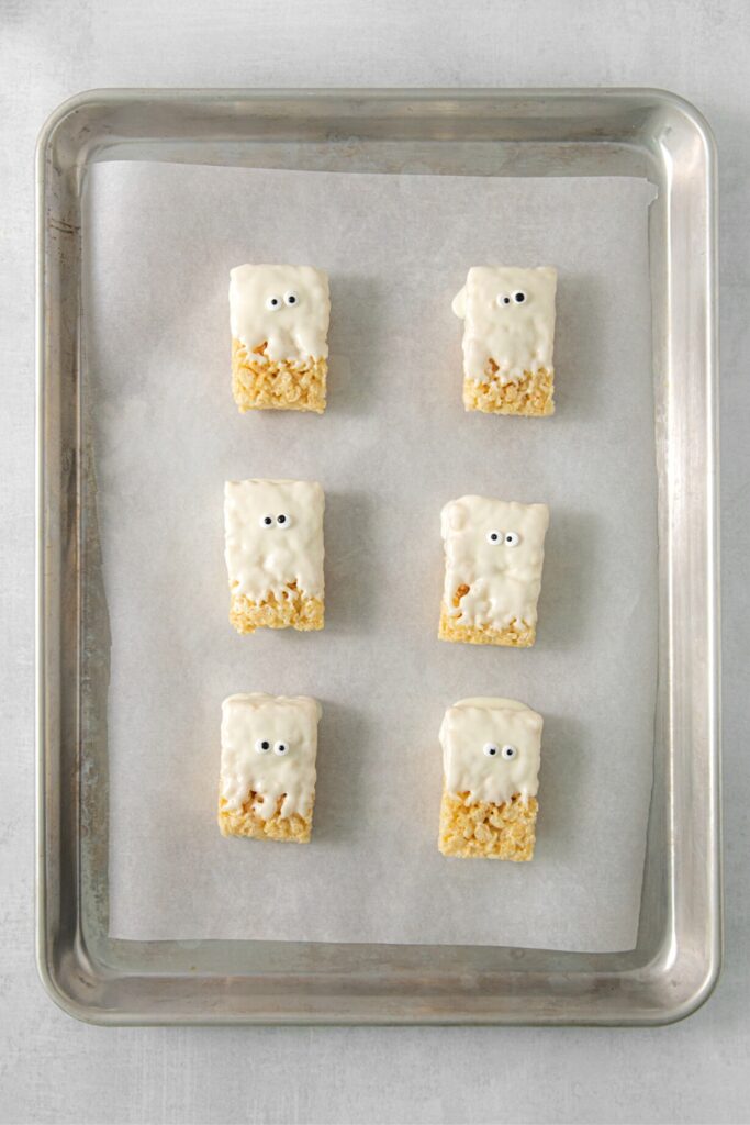Six mummy Rice Krispie Treats with candy eyes on a lined baking tray.