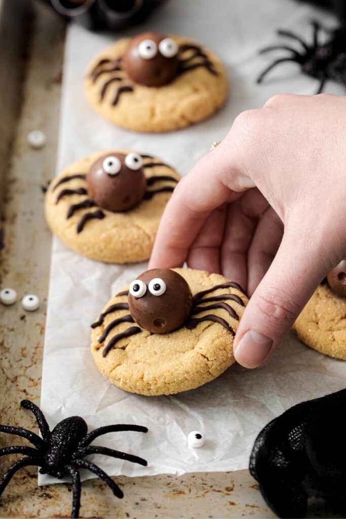 A hand lifting a peanut butter spider cookie off a lined baking tray.