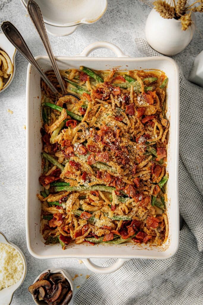 Cheesy green bean casserole with bacon in a white baking dish with 2 serving utensils.